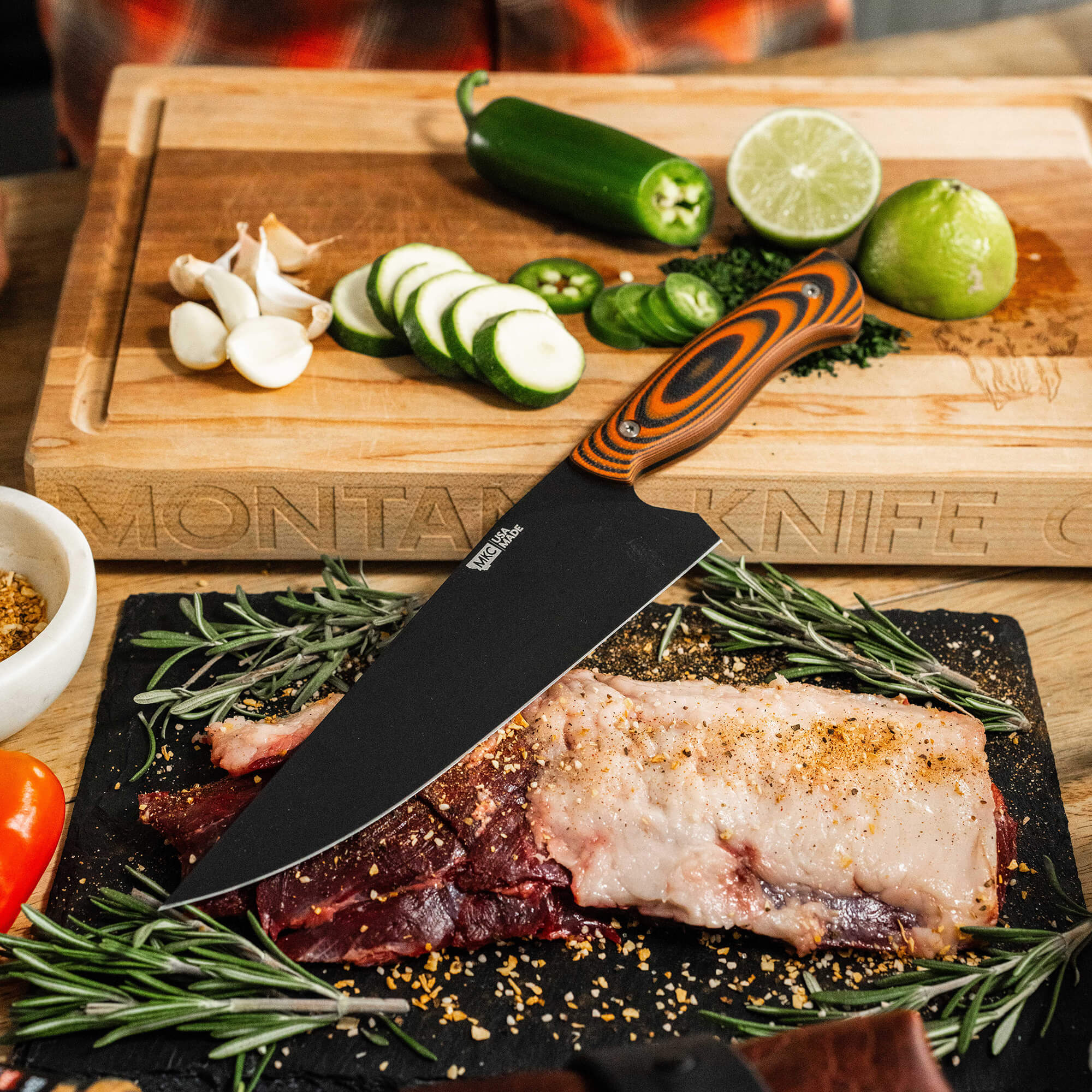 Alt text: "Bighorn Chef knife with a distinctive orange and black handle on a cutting board. The board features ingredients including sliced lime, cucumber, jalapeño, garlic cloves, and a raw seasoned steak surrounded by rosemary and spices,