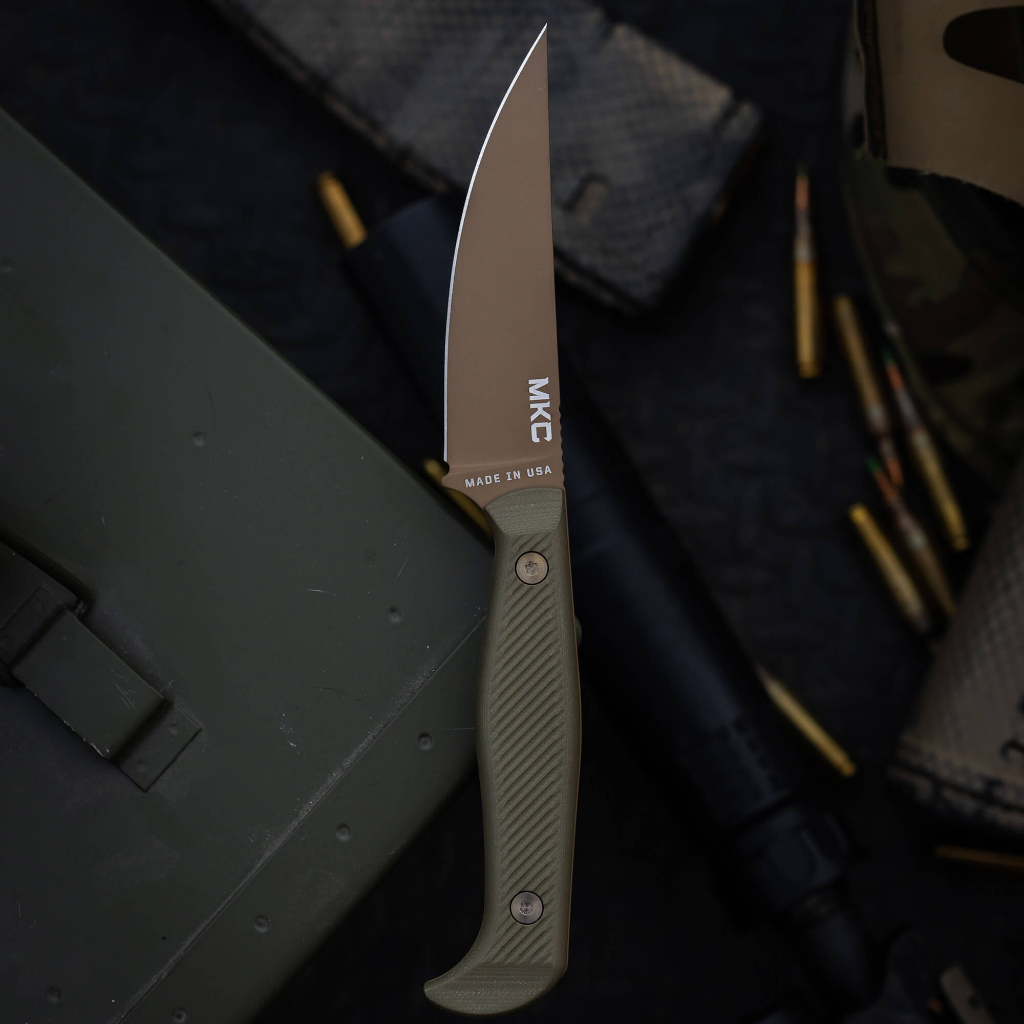 Alt text: Close-up of a MKC Battle Goat tactical knife with a coyote tan blade and olive drab handle, displayed on a green tactical gear background with scattered ammunition.