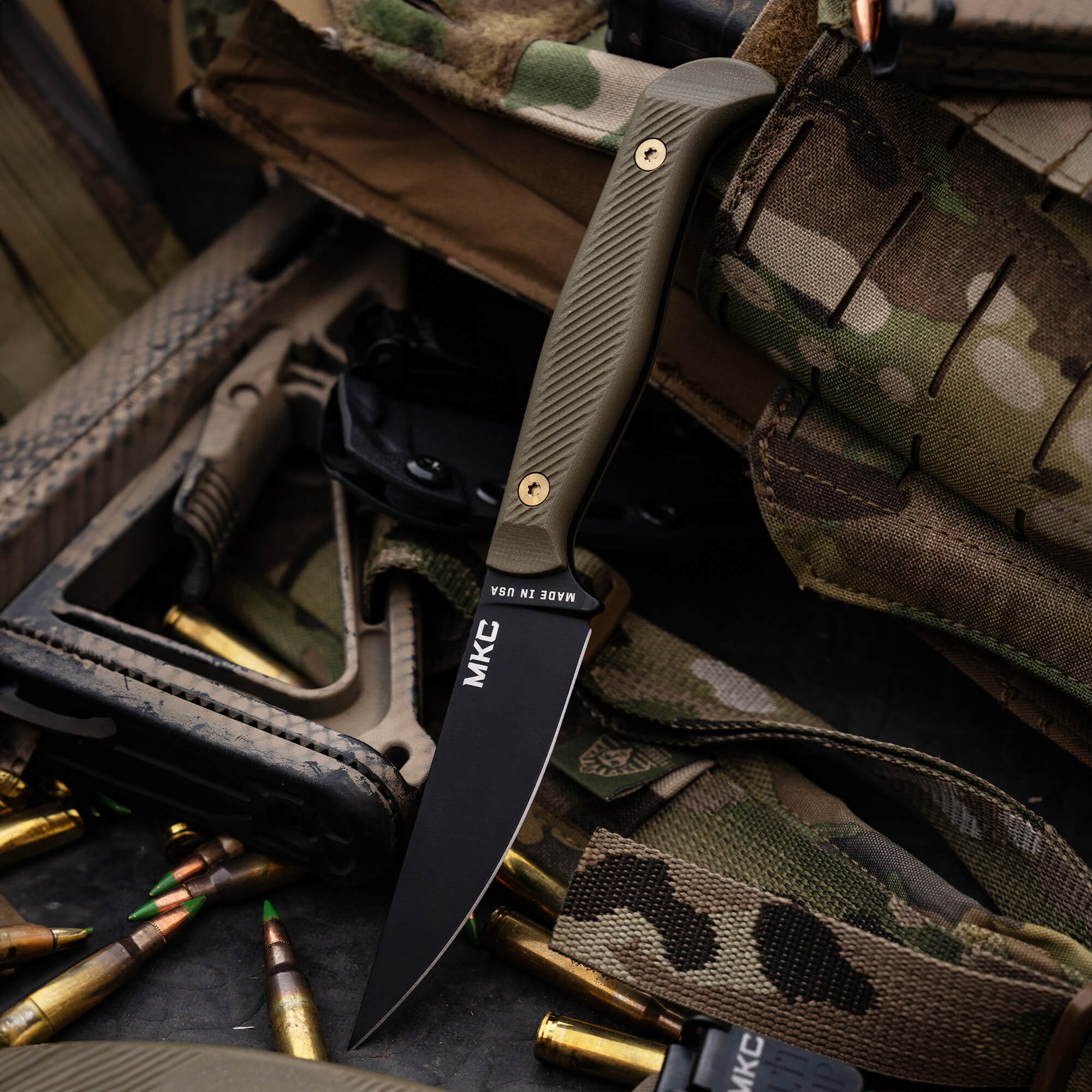 Alt text: A tactical folding knife with a black blade and olive green handle, labeled MKC, displayed atop a camouflage fabric background with scattered bullet casings and military gear.