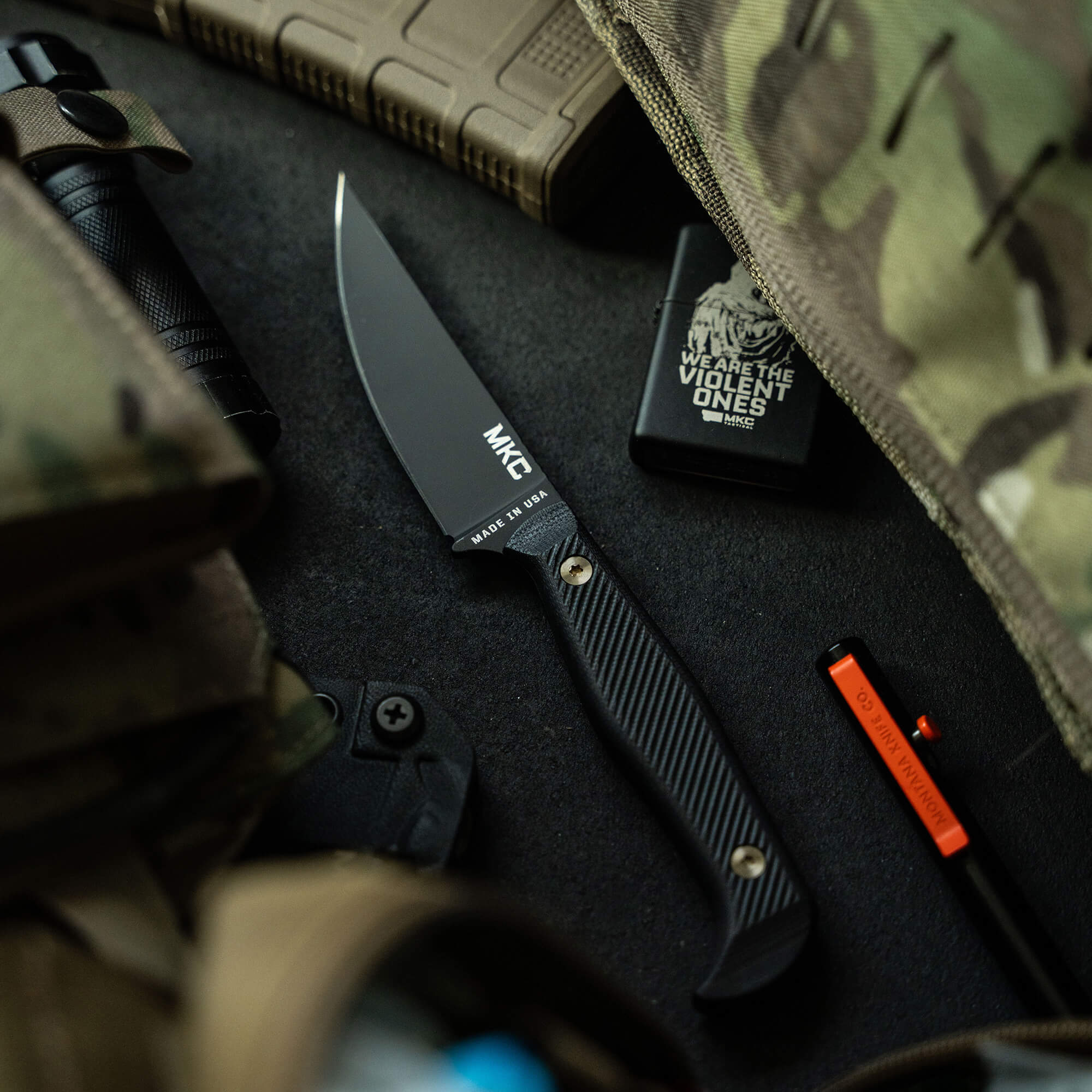 Alt text: "Black MKT fixed blade knife with textured handle displayed alongside tactical gear including a camouflage bag, lighter with a slogan, and a fire starter tool, set against a dark, rugged backdrop."