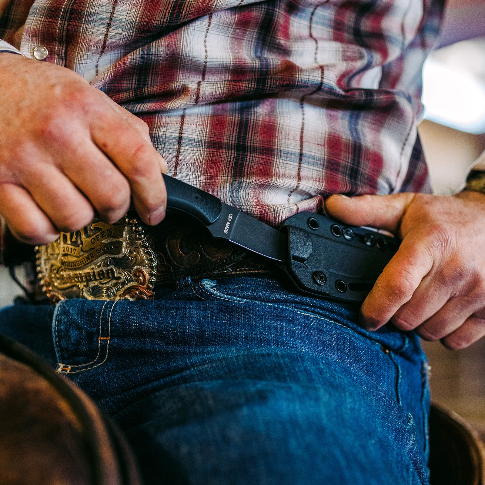 A man in a plaid shirt tucking a BLACKFOOT 2.0 knife in black color into his jeans pocket, with a detailed rodeo belt buckle visible.