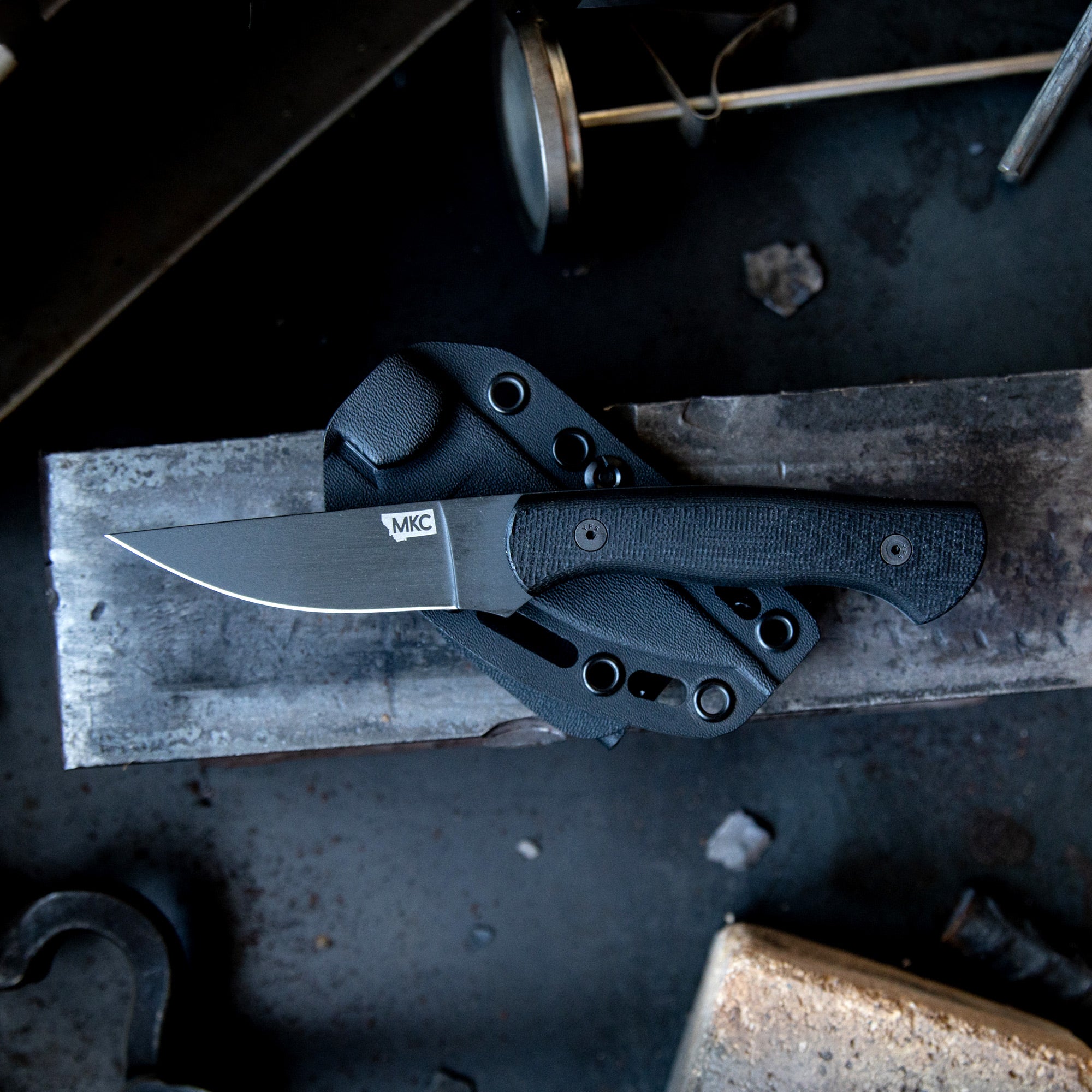 Alt text: "BLACKFOOT 2.0 tactical knife with a black handle and blade, displayed with its sheath on a gritty metal workshop surface, emphasizing its rugged design and durability."
