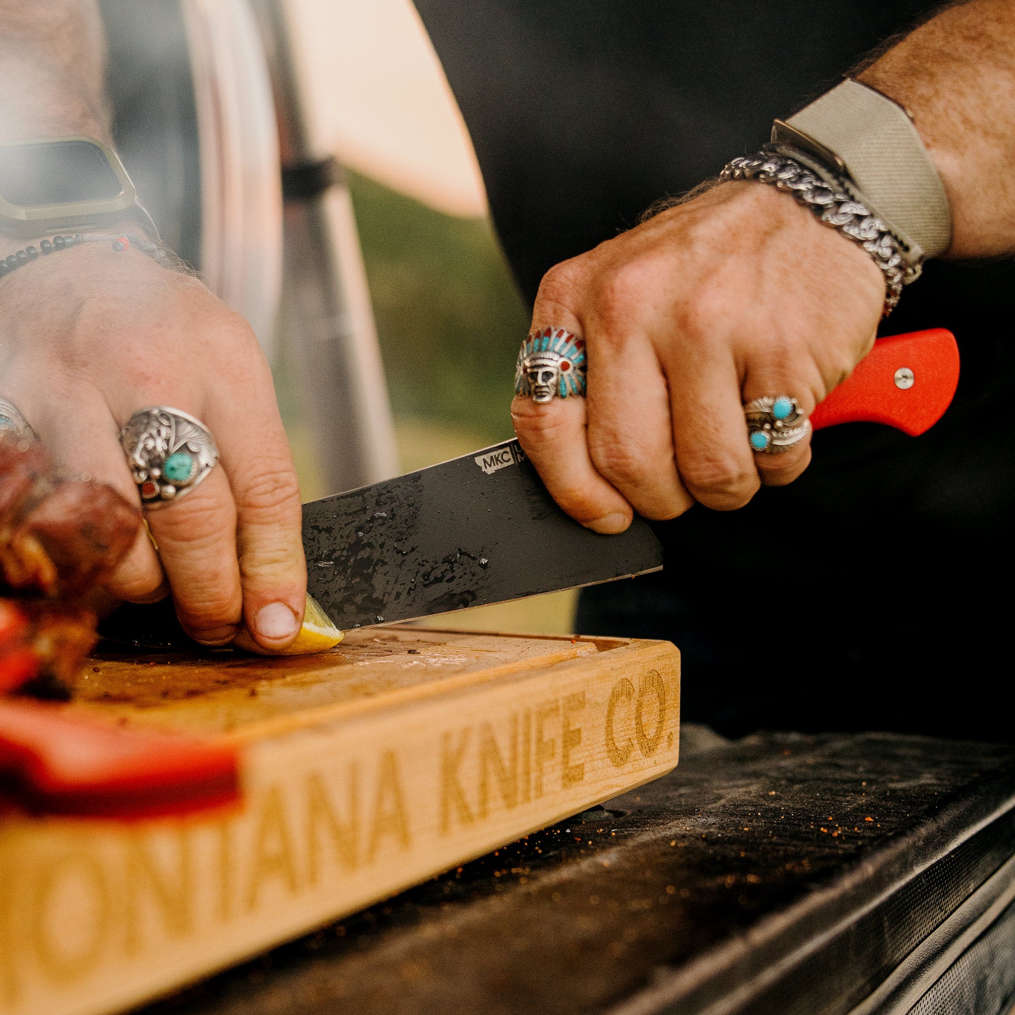 Close-up of a person's hands wearing decorative rings, slicing meat on a wooden cutting board with a Bighorn Chef knife featuring a red handle, marked by Montana Knife Co. logo.