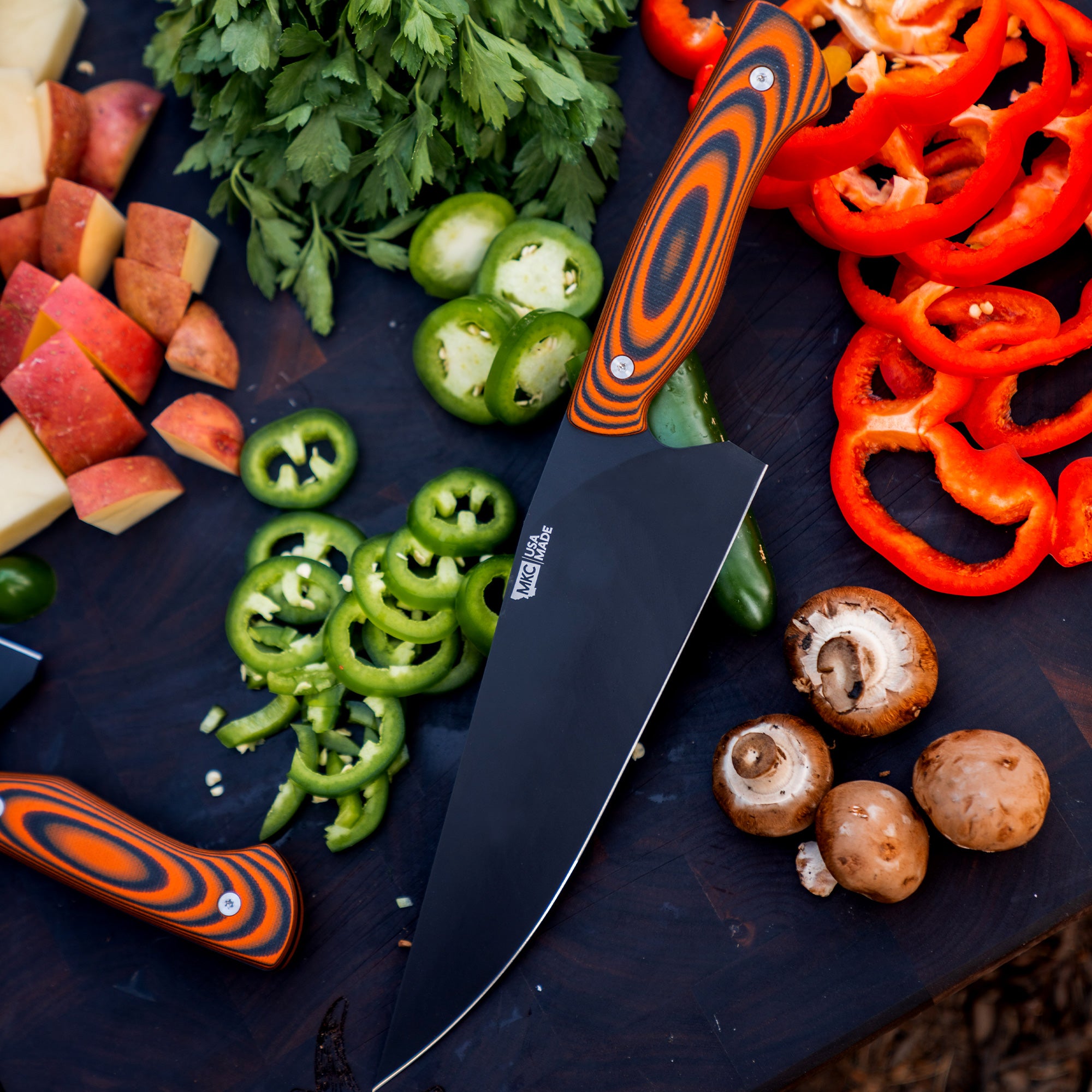 Orange and black handled Bighorn Chef knife on a wooden surface surrounded by freshly cut vegetables including red and green bell peppers, mushrooms, and assorted fruits.