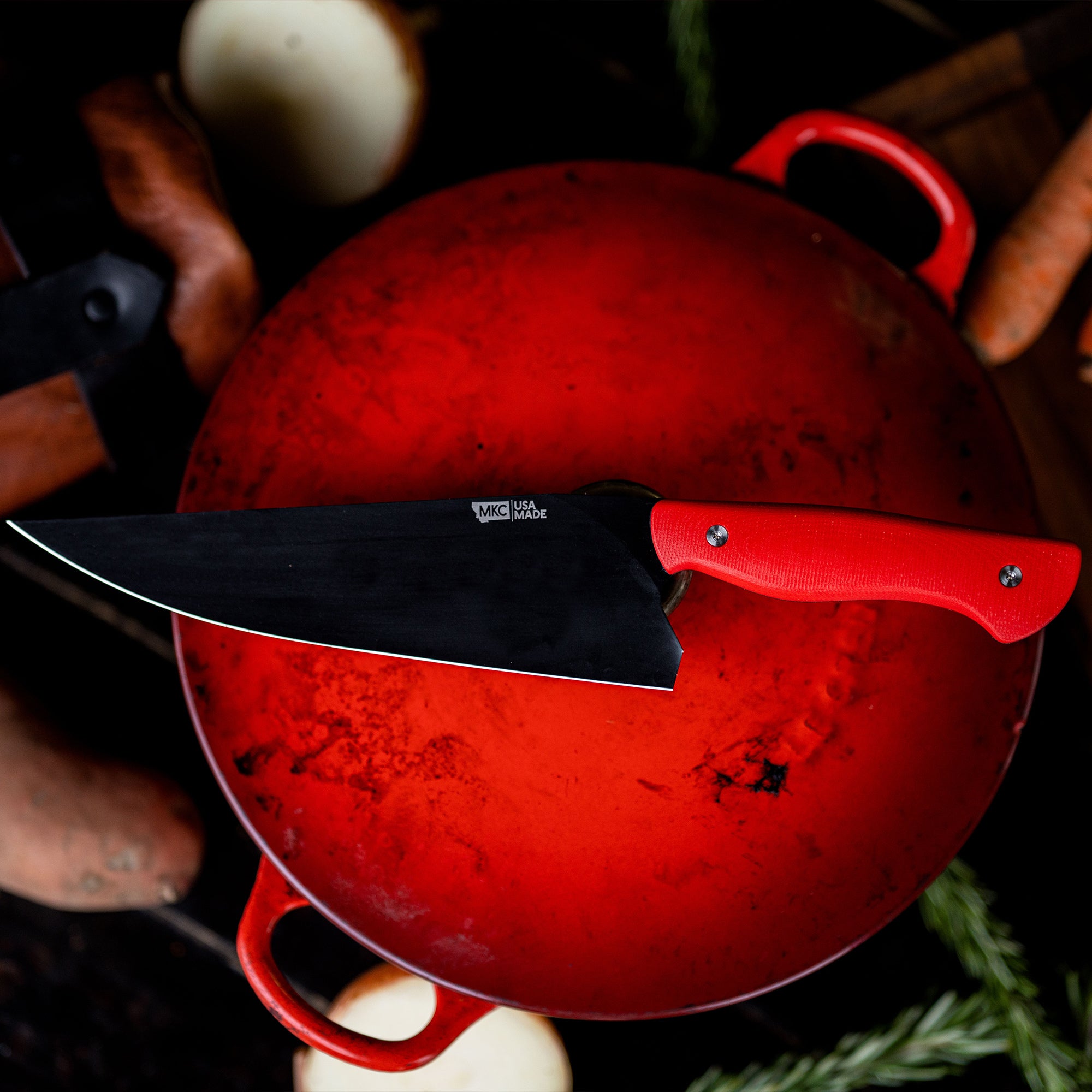 Alt text: "A vibrant red Bighorn Chef knife with a matte black blade, resting on a matching red enameled cast iron pot, surrounded by rustic kitchen ingredients like onions and carrots."