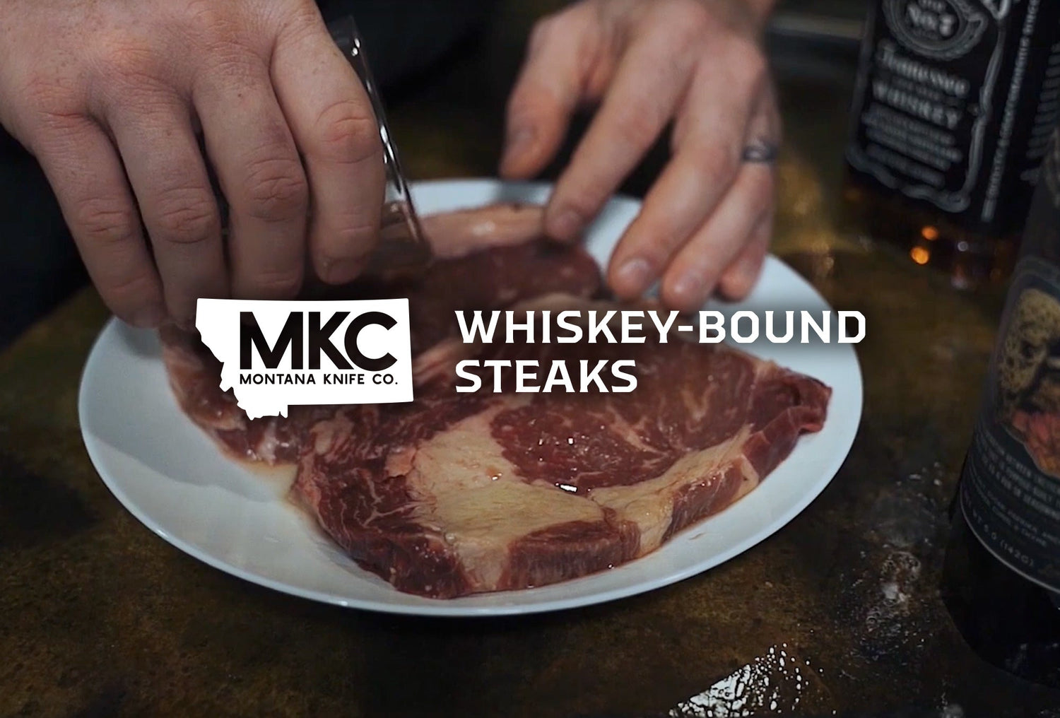 Hands using a knife to prepare raw ribeye steaks on a white plate with whiskey bottle in the background.