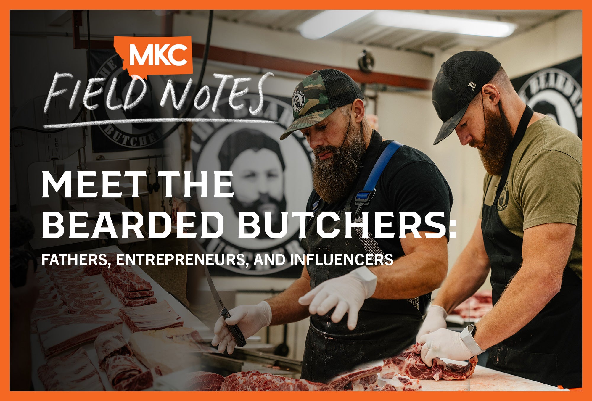 Two bearded butchers wearing aprons and caps work together cutting meat at a butcher block counter in a commercial kitchen.