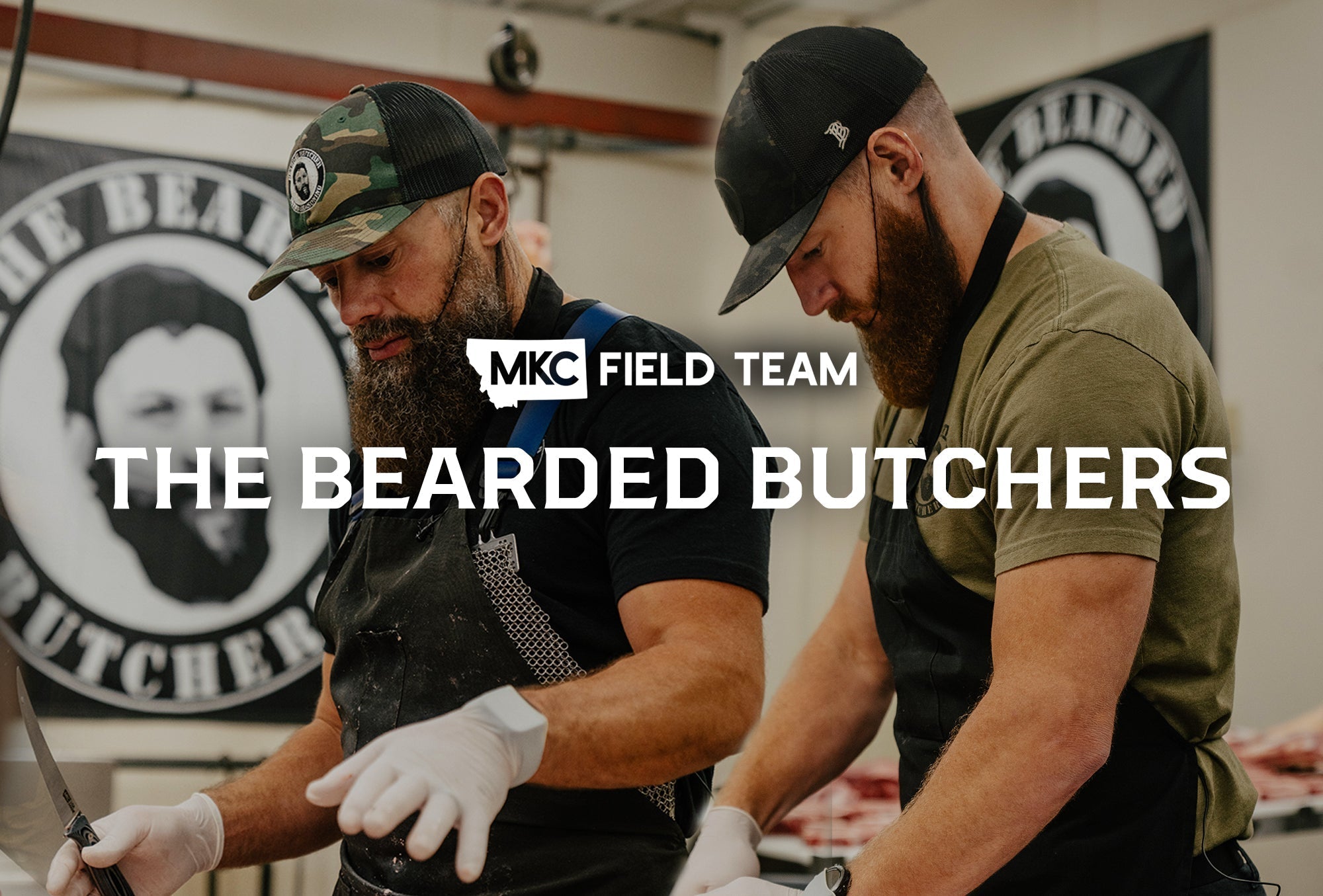 Two bearded butchers wearing aprons and caps work together cutting meat at a butcher block counter in a commercial kitchen.