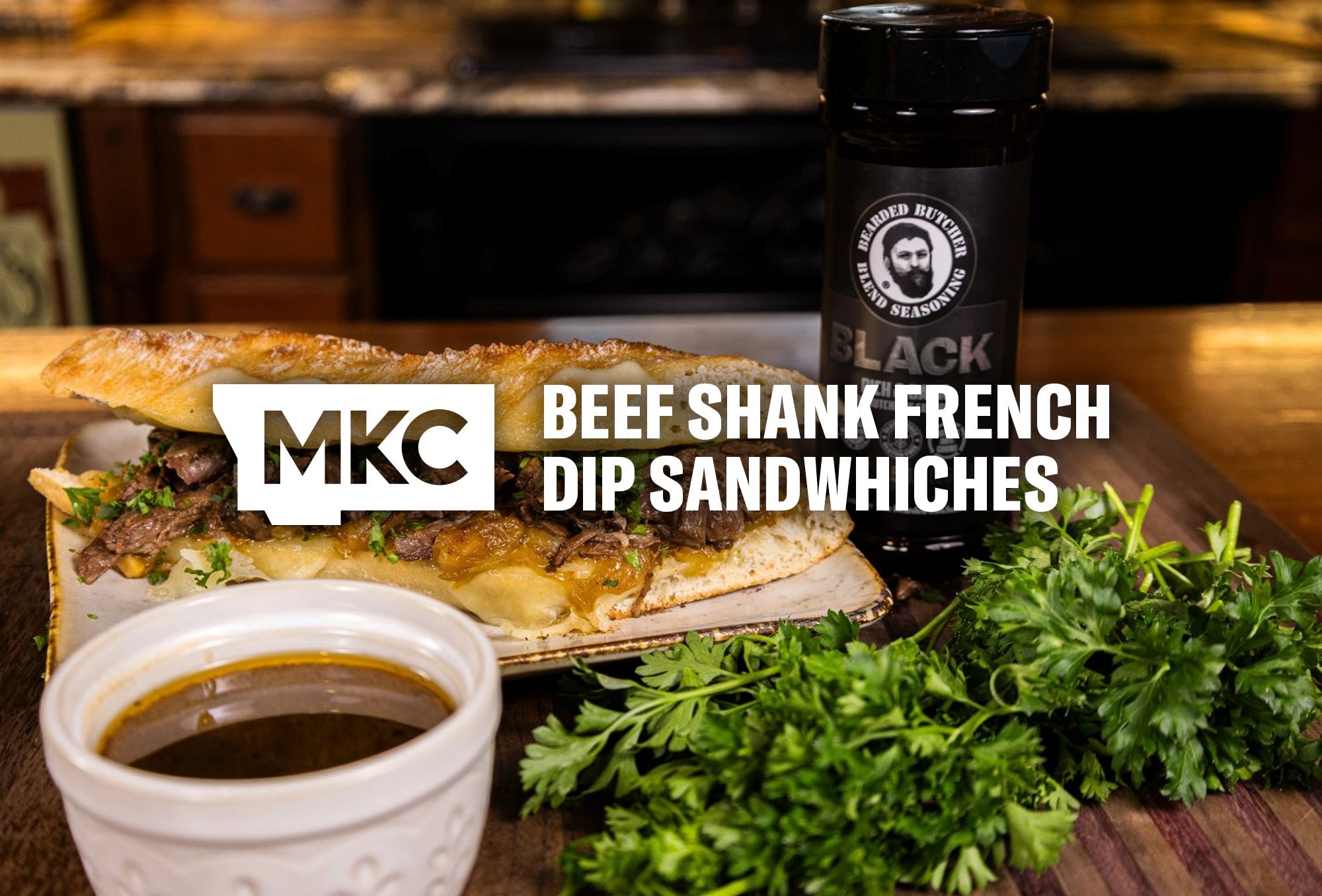 A braised beef shank sandwich on a table beside fresh herbs, seasoning, and a bowl of jus.