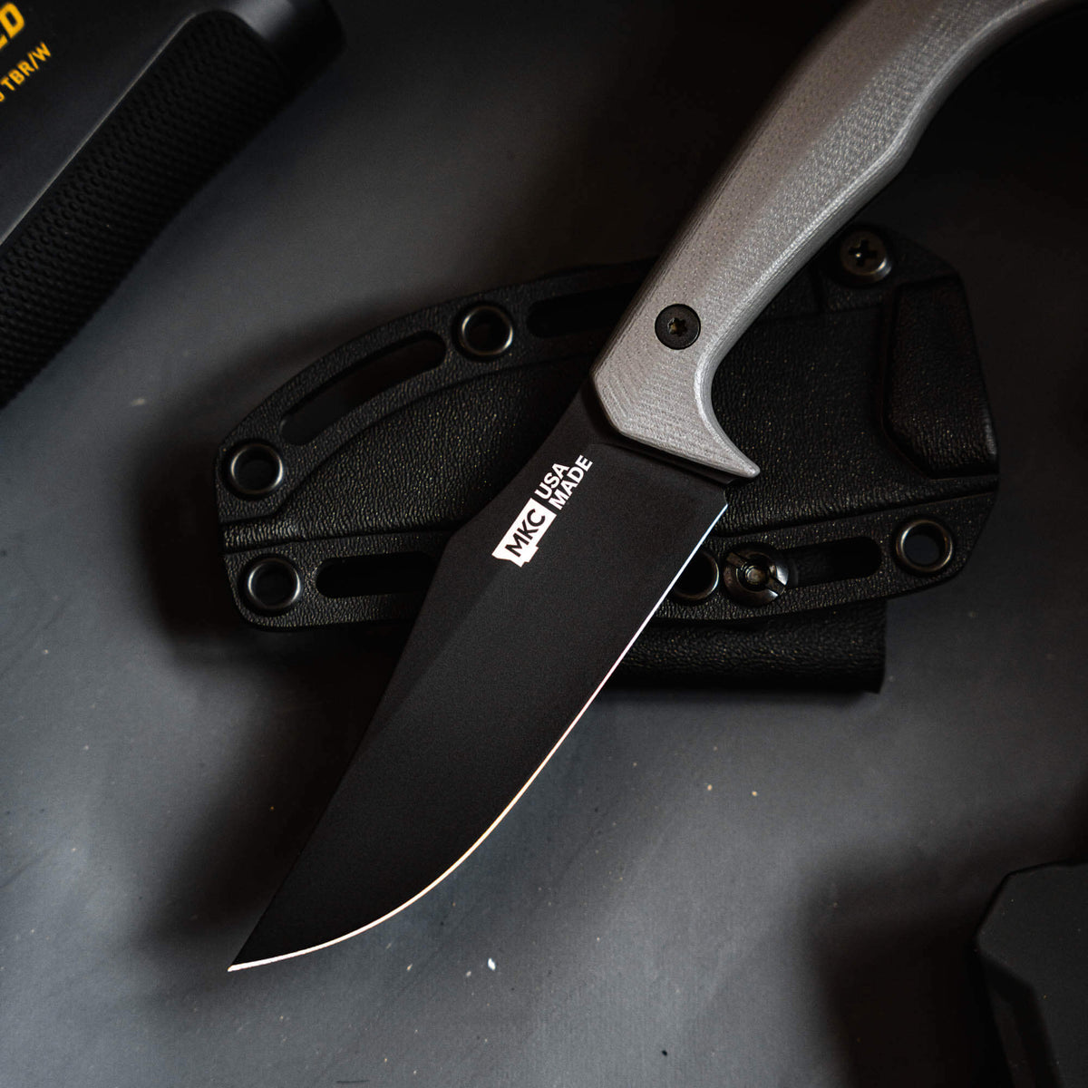 Grey Triumph Pro knife with a black blade resting on a textured black sheath, showcasing its sleek design and functionality.