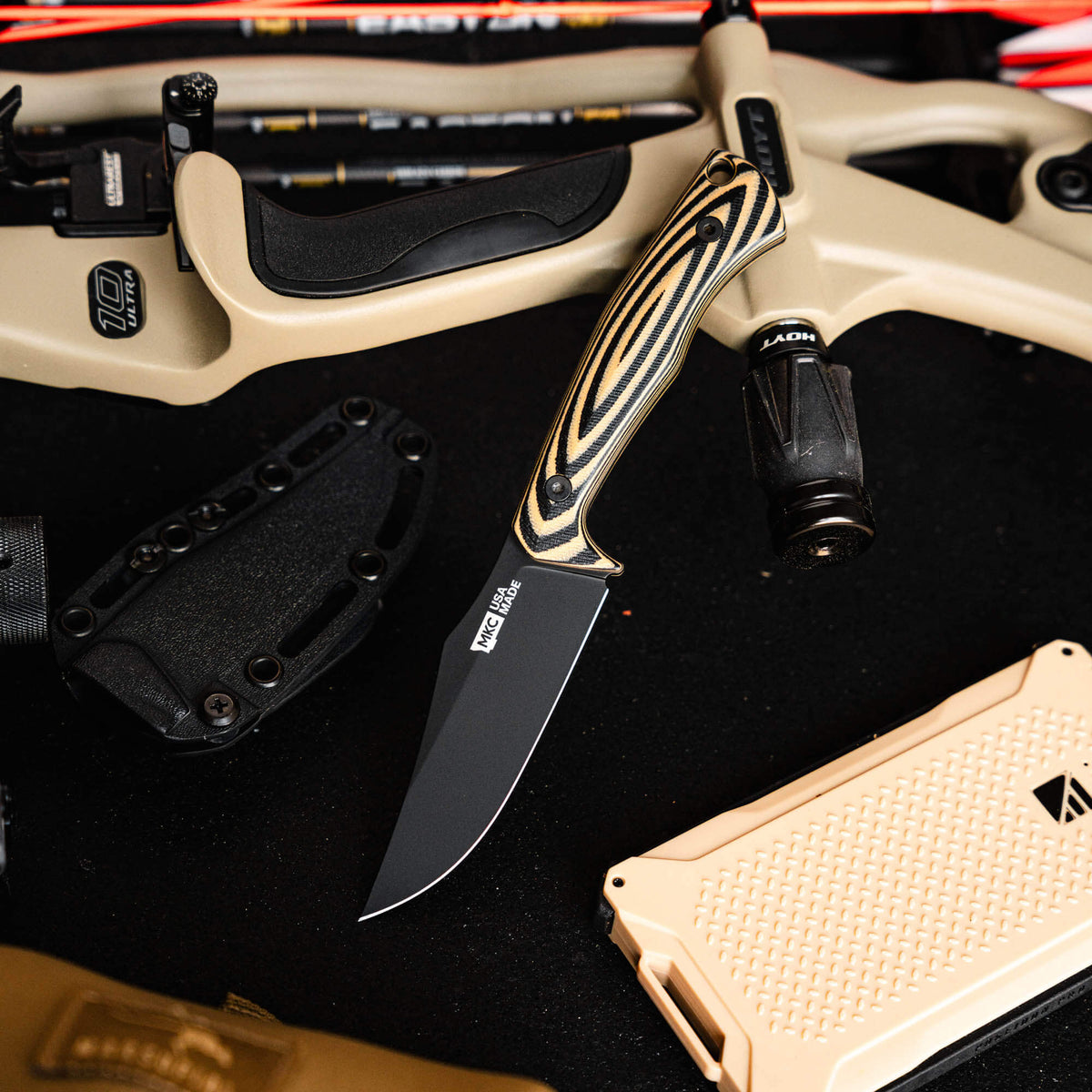 Triumph Pro knife in tan and black with a sleek blade, surrounded by outdoor gear and equipment.