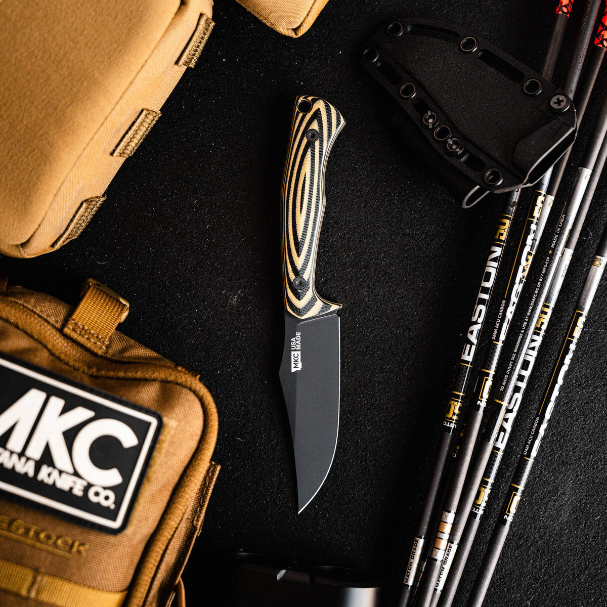Triumph Pro knife in tan and black with a sleek blade, surrounded by outdoor gear and accessories on a dark surface.
