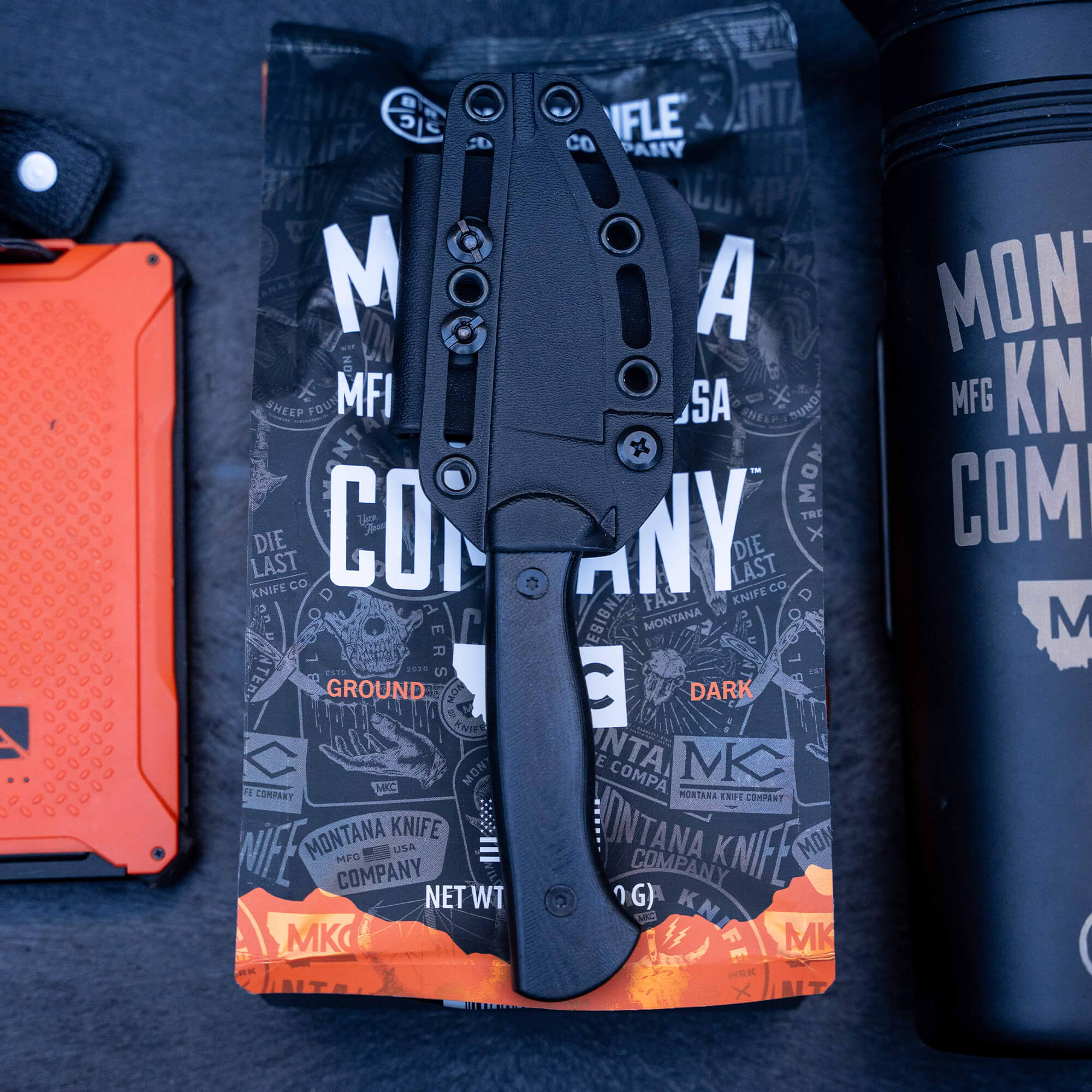 Alt text: "Close-up of a BLACKFOOT 2.0 tactical knife in black color, sheathed and attached to a Kydex holster, displayed on a themed packaging backdrop with coffee beans and a survival water bottle."