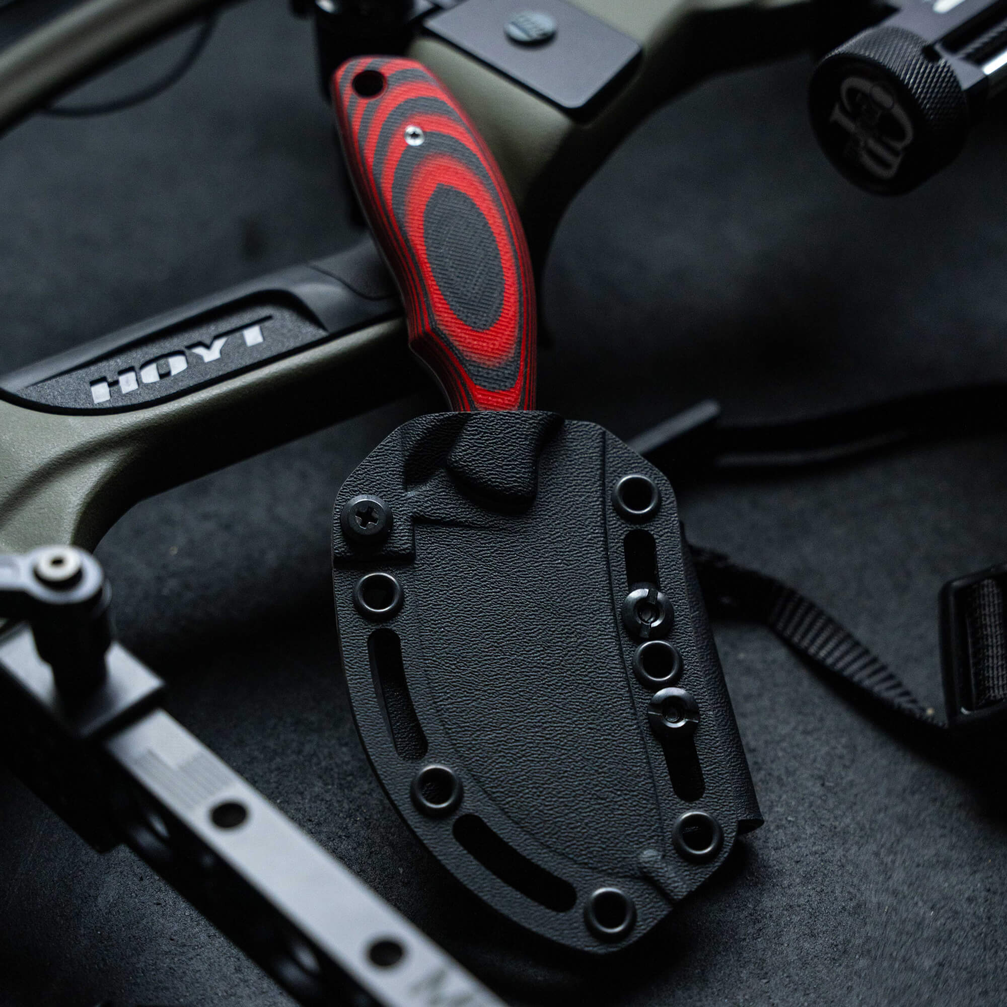 Alt text: "Close-up of a red and black handled skinner knife in a black sheath, displayed on a dark textured surface with archery equipment in the background."