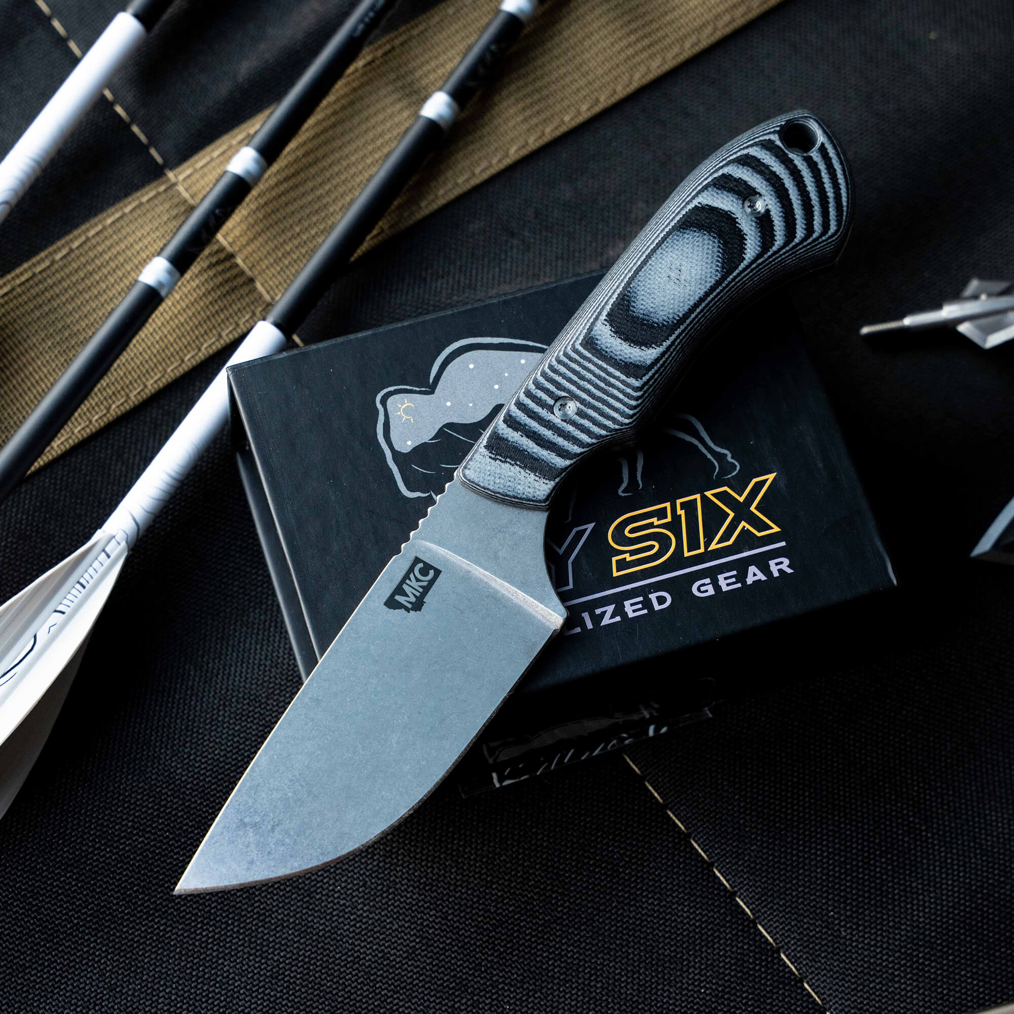 MKC Elk Knife with a sleek blade and textured handle, displayed on a black box with fishing gear in the background.