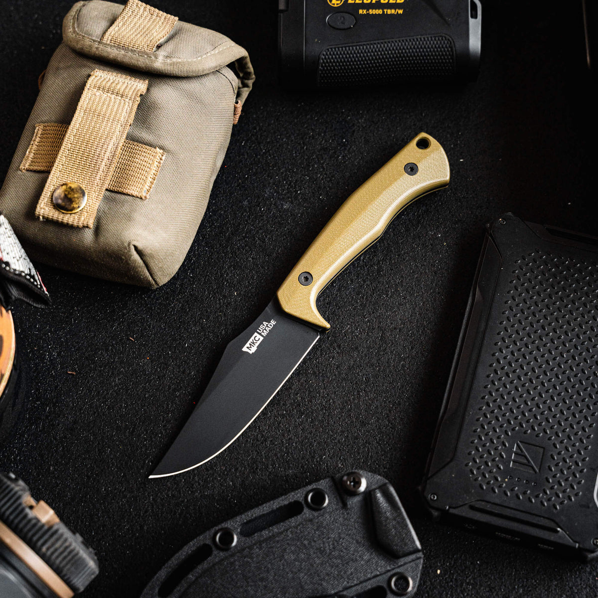 Olive green Triumph Pro knife with a black blade, surrounded by tactical gear and accessories on a dark surface.