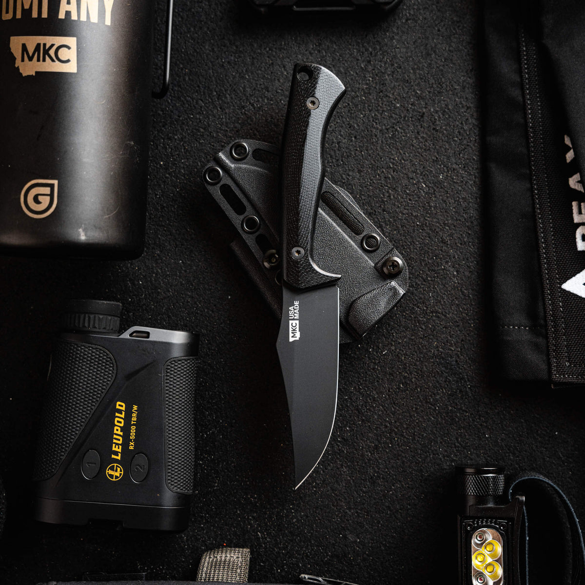 Triumph Pro Black knife displayed on a textured surface with outdoor gear, showcasing its sleek design and functionality.