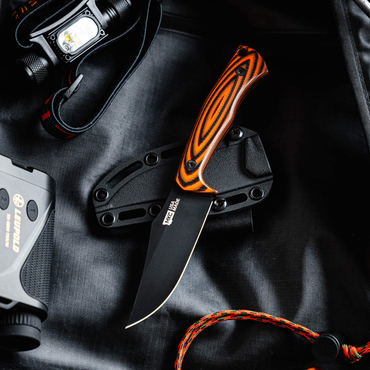 Triumph Pro knife with orange and black handle, black blade, and sheath, placed on a dark background with gear.