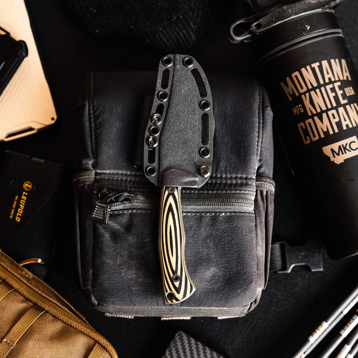Triumph Pro knife in tan and black with a sleek handle, resting on a dark bag among outdoor gear and tools.