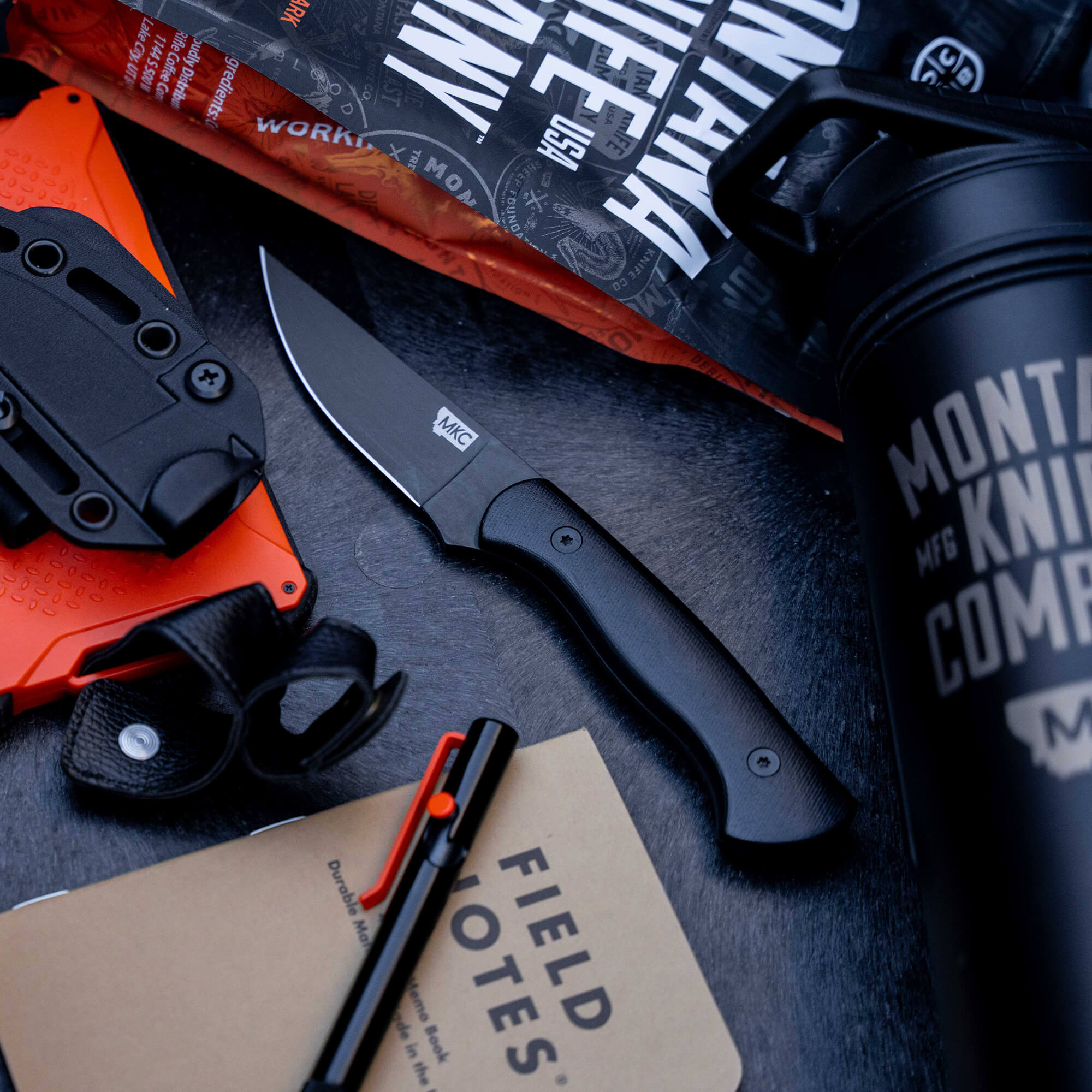 Alt text: "BLACKFOOT 2.0 tactical knife displayed with accessories including a sheath, pen, notebook, and a water bottle, all arranged on a textured dark surface."