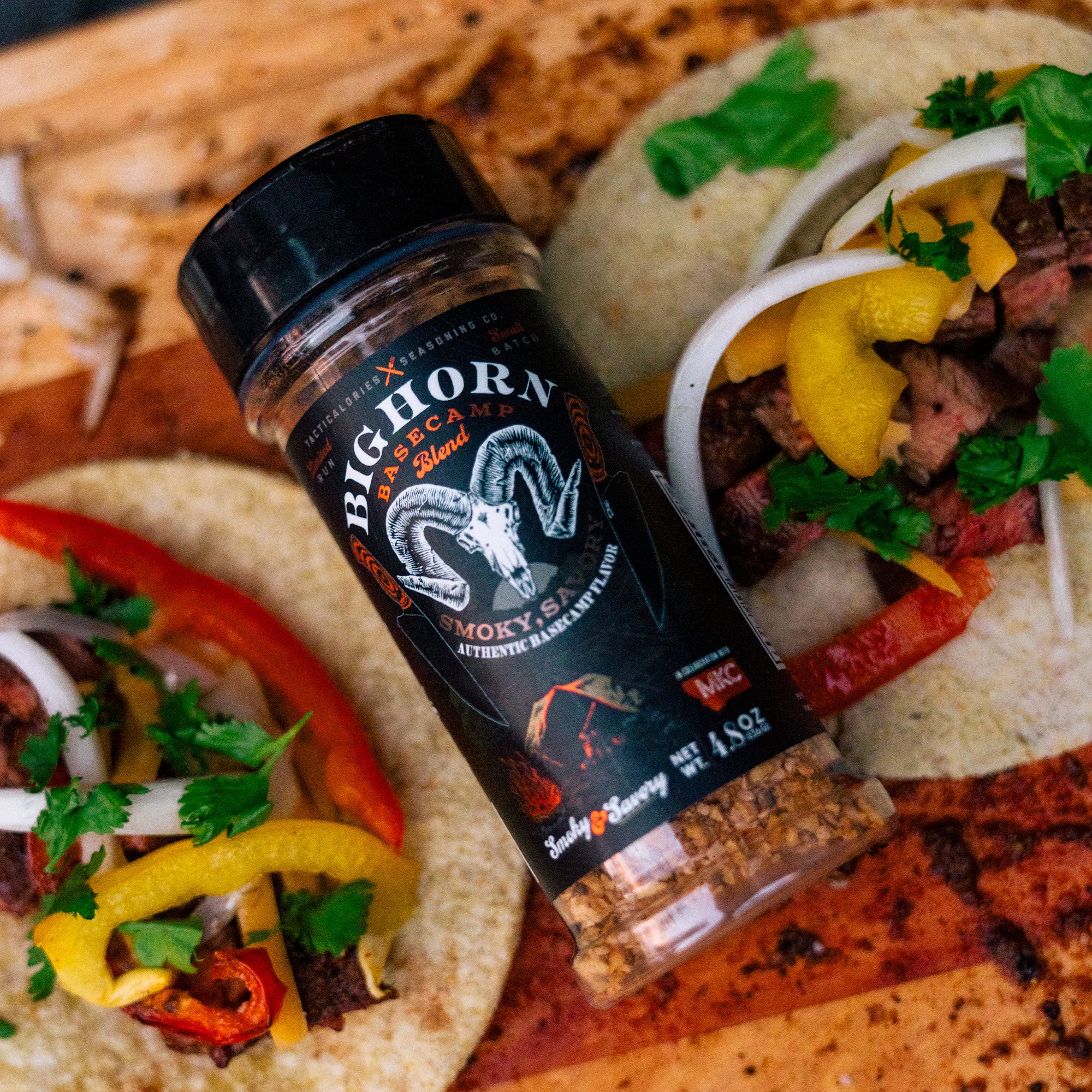 Alt text: "A bottle of Bighorn Basecamp Seasoning on a wooden surface, labeled 'Smoky & Savory' with an image of a ram's head, displayed alongside tacos with beef, fresh cilantro, onions, and sliced