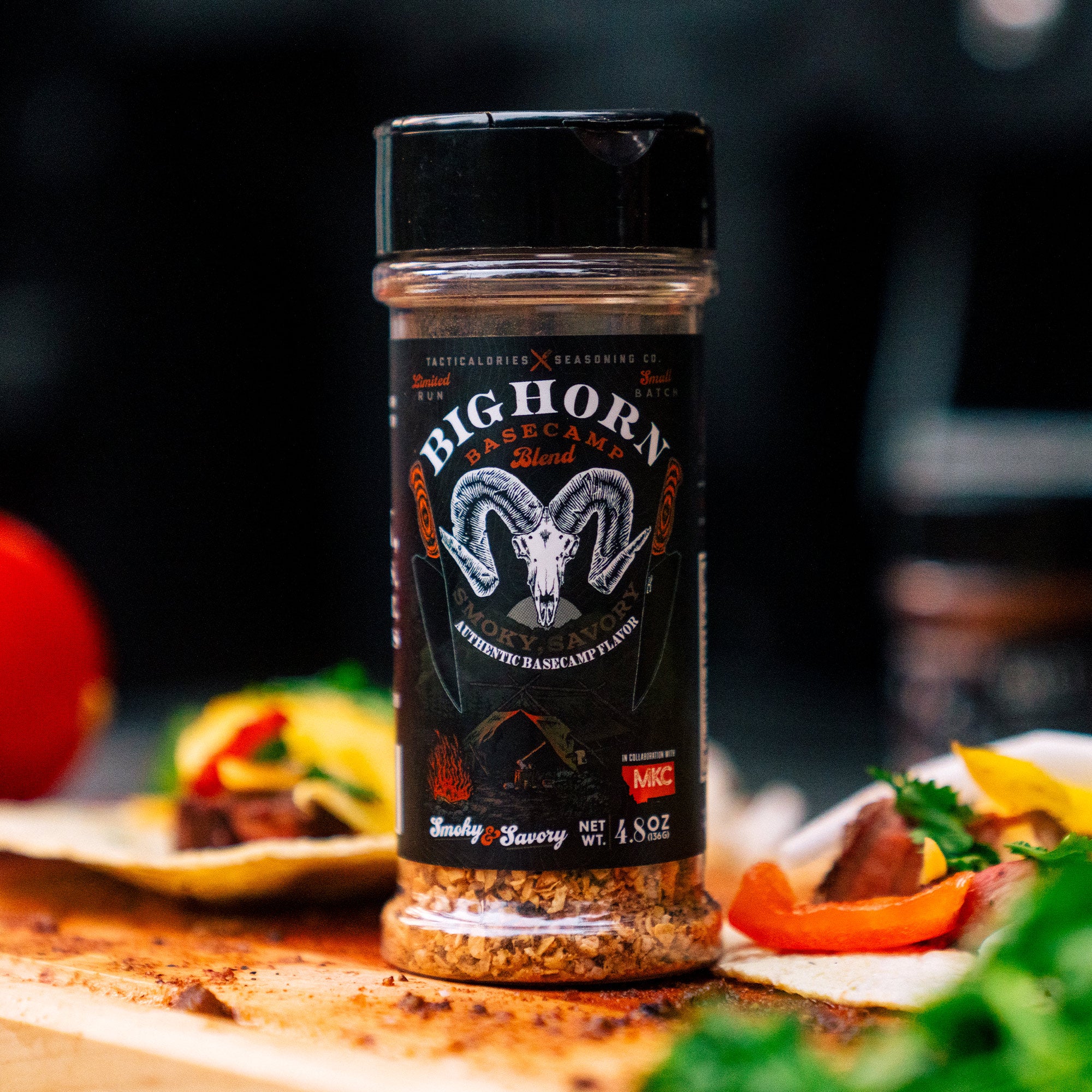 Alt text: "BIGHORN Basecamp Seasoning bottle with a clear view of its label featuring a white ram skull, campfire graphic, and text 'Smoky & Savory'. The bottle is placed on a wooden surface surrounded by food ingredients