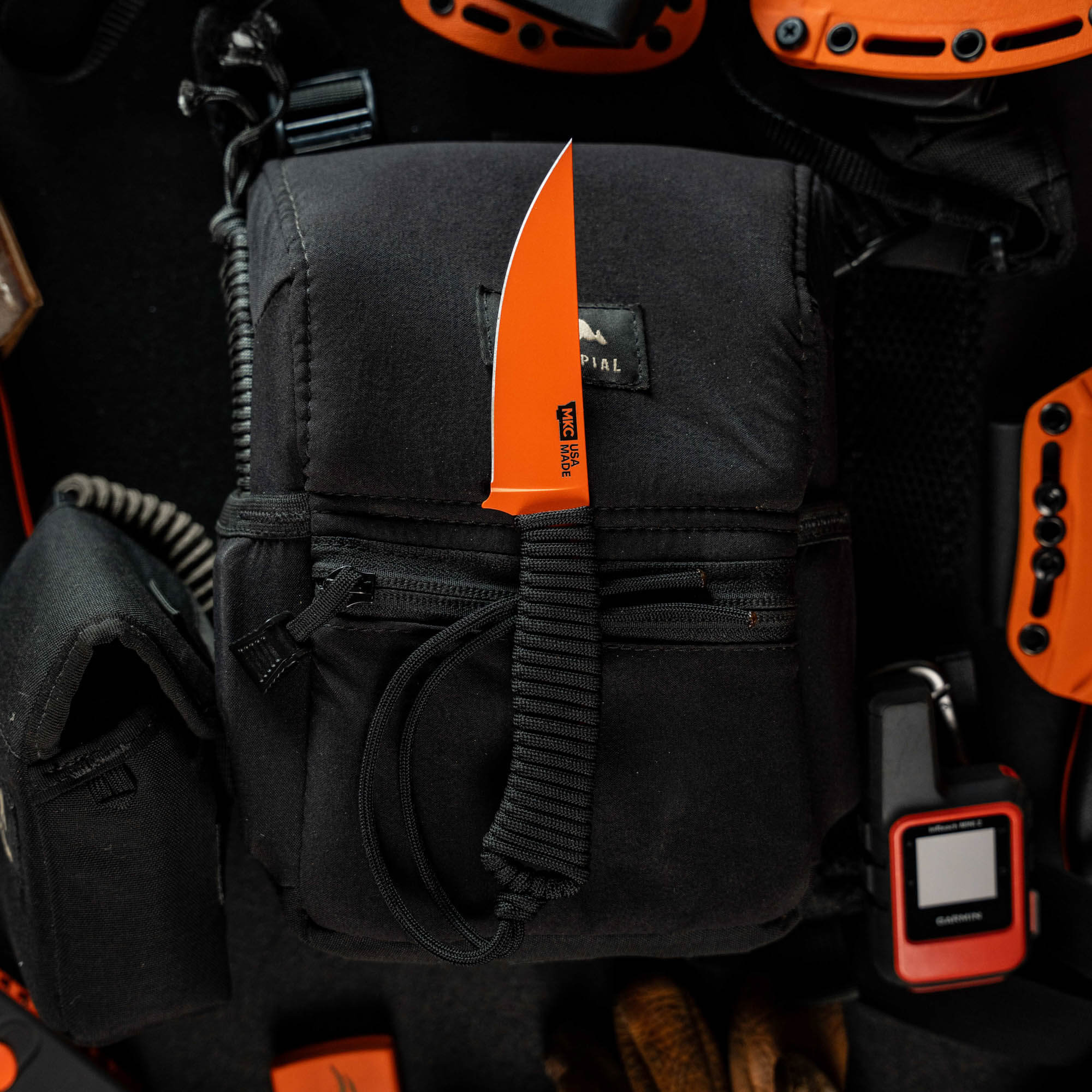 SPEEDGOAT 2.0 knife with blaze orange Cerakote finish resting on a black tactical pouch, surrounded by outdoor gear.