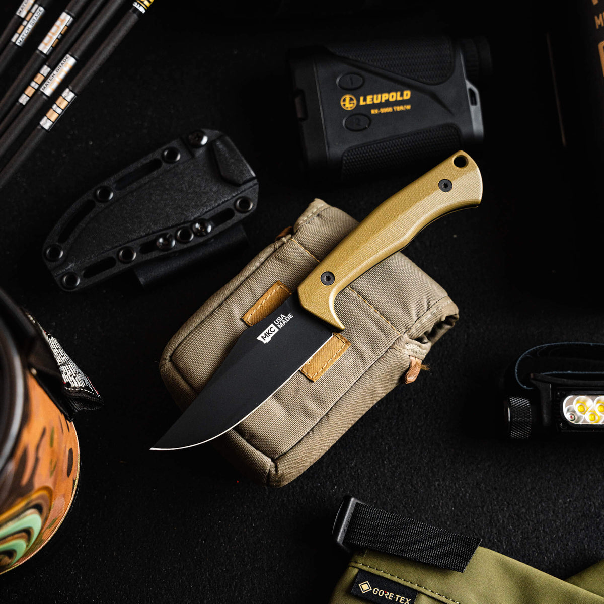 Olive TRIUMPH PRO knife on a pouch, surrounded by outdoor gear, showcasing its rugged design for hunters and outdoorsmen.