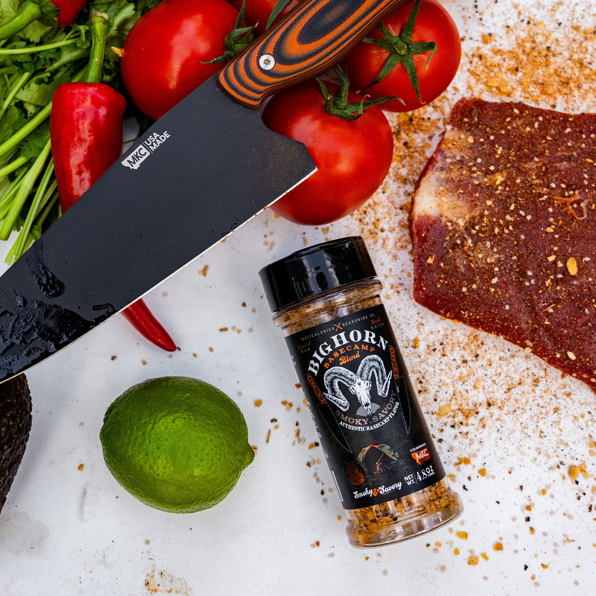 Alt text: "A bottle of Bighorn Basecamp Smoky Sassy Seasoning next to fresh ingredients including tomatoes on the vine, a lime, and a dusting of spices on a white surface, with a large kitchen knife partially covering the