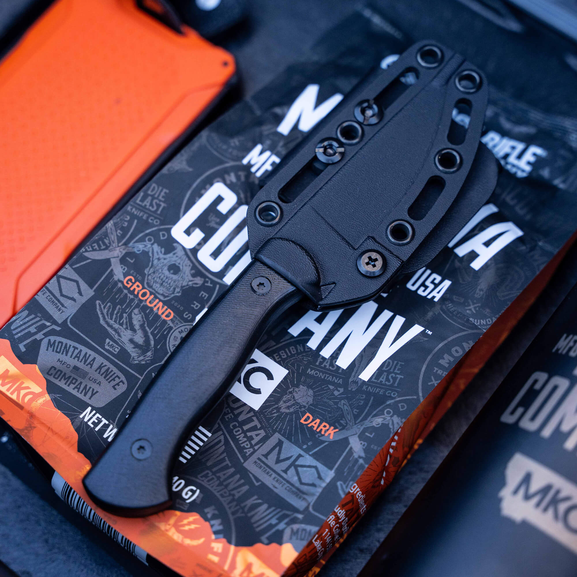 Alt text: Black BLACKFOOT 2.0 tactical knife in a matte finish, displayed partially unsheathed from a black holster, resting on colorful packaging with text and graphics from Montana Knife Company.