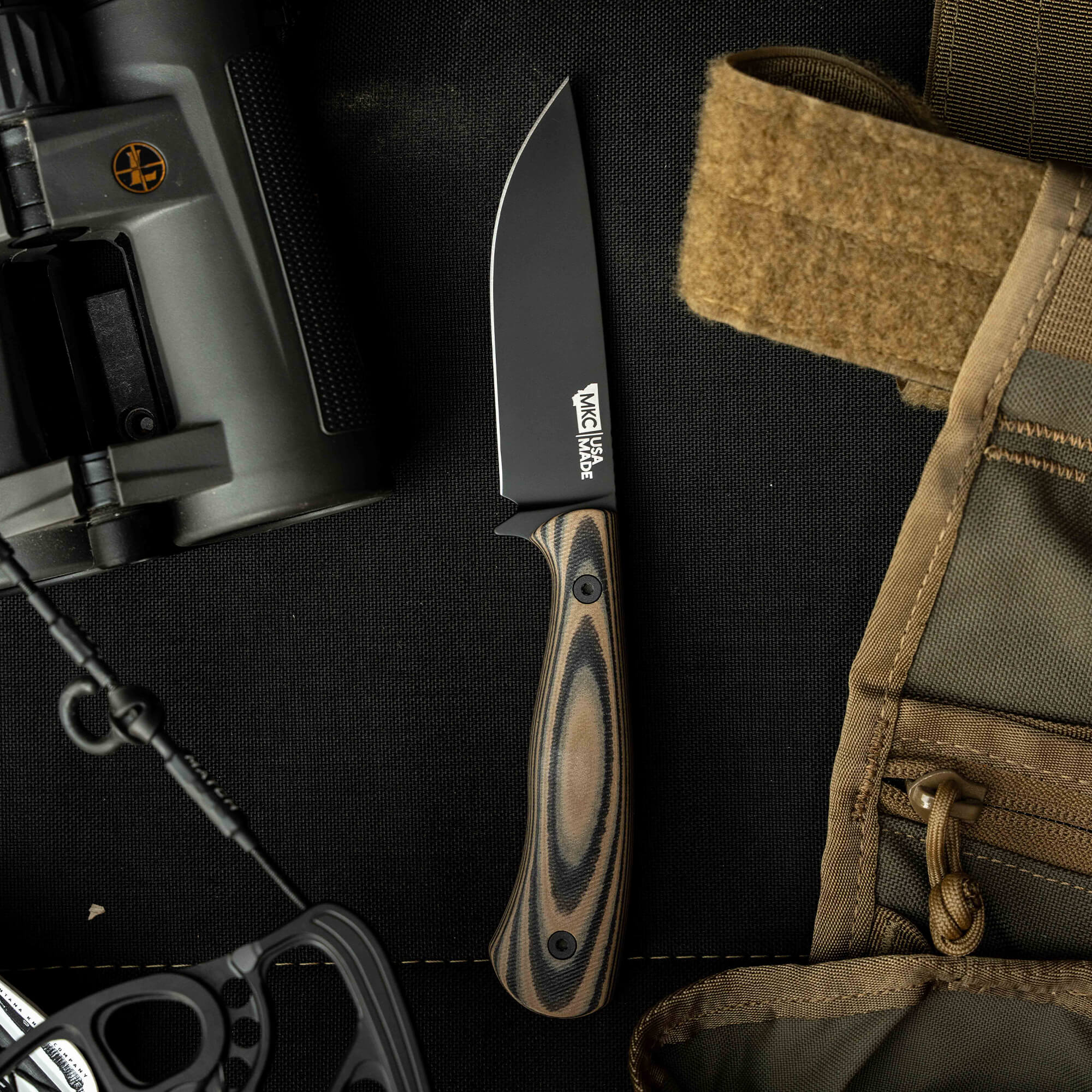 Mathews Archery MKC Whitetail knife with a black blade and patterned handle, surrounded by outdoor gear.