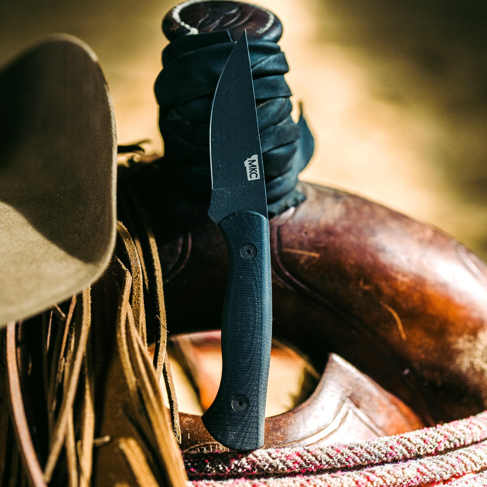 Alt text: "Close-up image of the BLACKFOOT 2.0 tactical knife in black, displayed with its blade partially sheathed in a leather wrap, resting against a rustic leather saddle and fringe. The focus is on the textured handle and