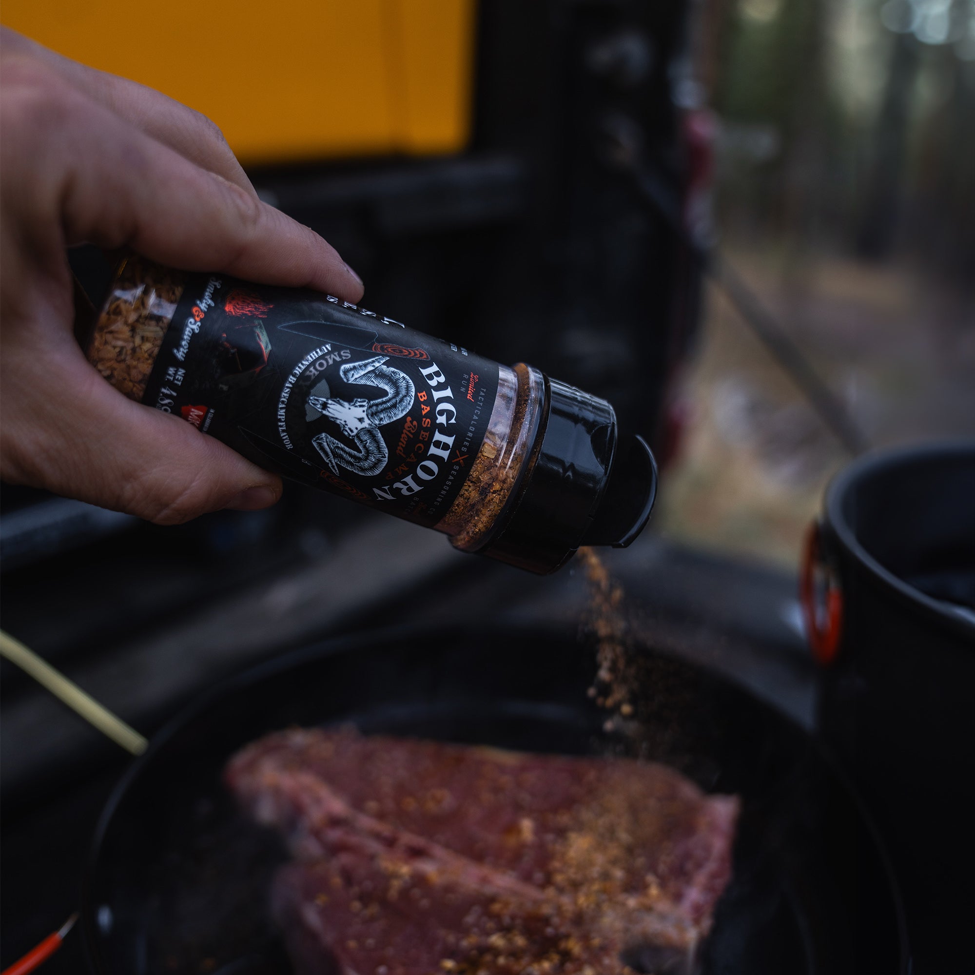 Hand applying Bighorn Basecamp Seasoning from a black bottle to meat on a skillet, set against a blurred outdoor camping background.