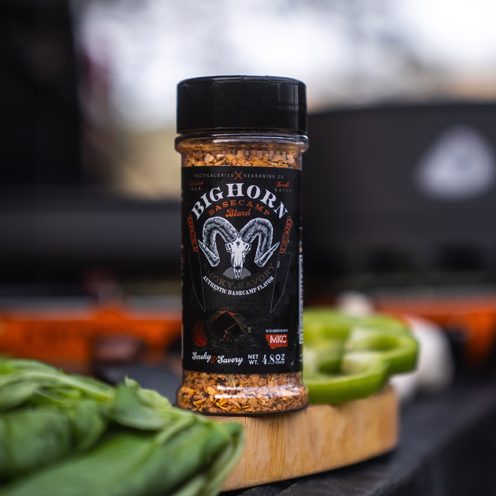 Close-up of a Bighorn Basecamp Seasoning bottle on a wooden cutting board, prominently displaying the label and spices inside, with blurred fresh greens and a cooking setup in the background.