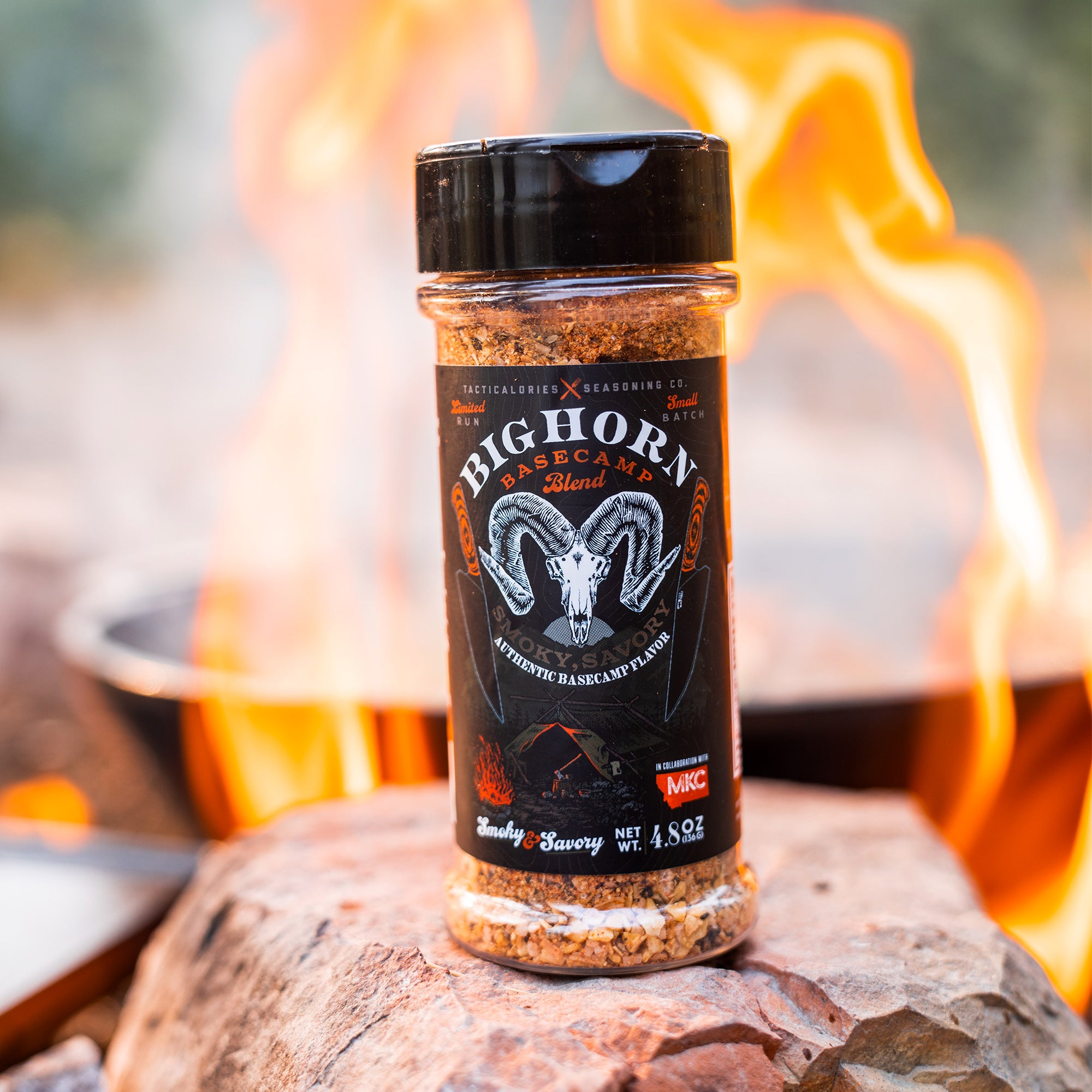 Alt text: "Bighorn Basecamp Blend seasoning bottle by Tacticalories Seasoning Co., prominently displayed on a rocky surface with a campfire blazing in the background, highlighting its smokey and savory flavor profile, perfect for outdoor cooking."