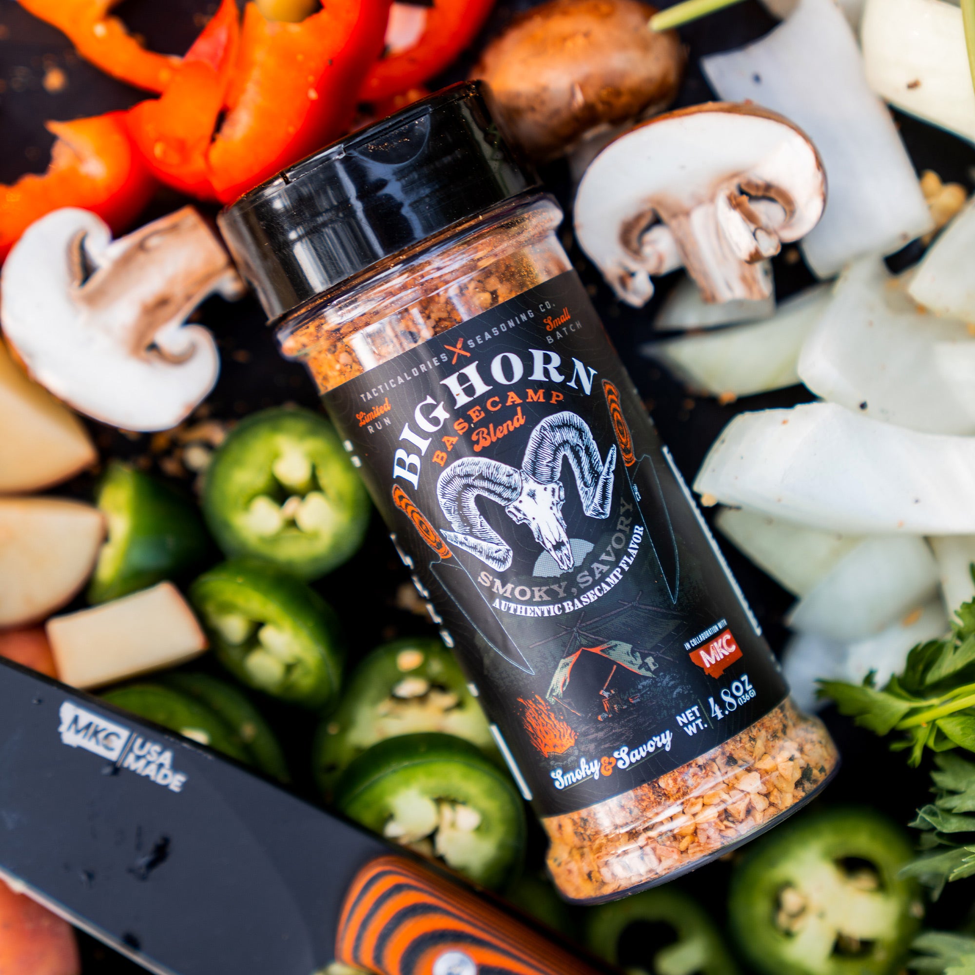 Alt text: "Bighorn Basecamp gourmet seasoning bottle labeled 'Smoky & Savory,' surrounded by vibrant, fresh ingredients including sliced red bell peppers, mushrooms, onions, and jalapenos."