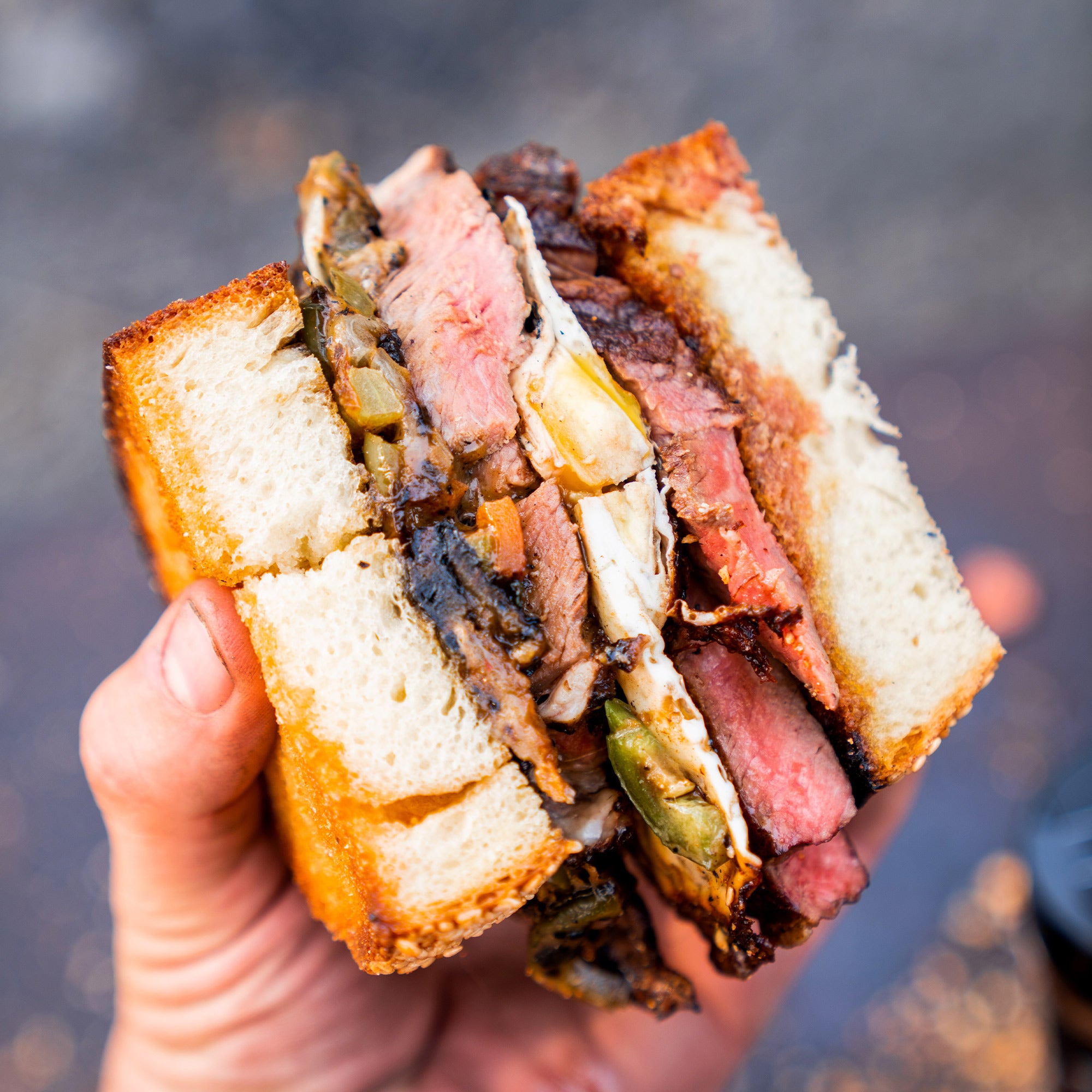 A close-up image of a hand holding a hearty sandwich filled with seasoned beef, vegetables, and melted cheese, highlighting the use of Bighorn Basecamp Seasoning.